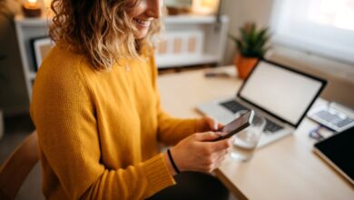 Beautiful young woman sitting at the desk in a home office and using a smart phone.