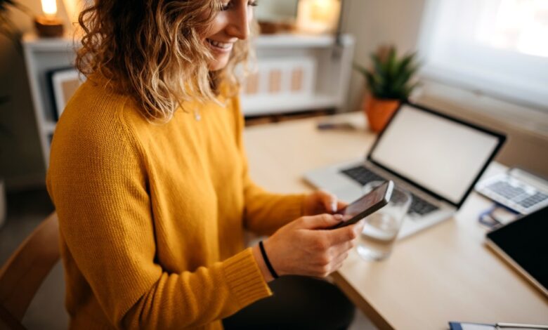 Beautiful young woman sitting at the desk in a home office and using a smart phone.