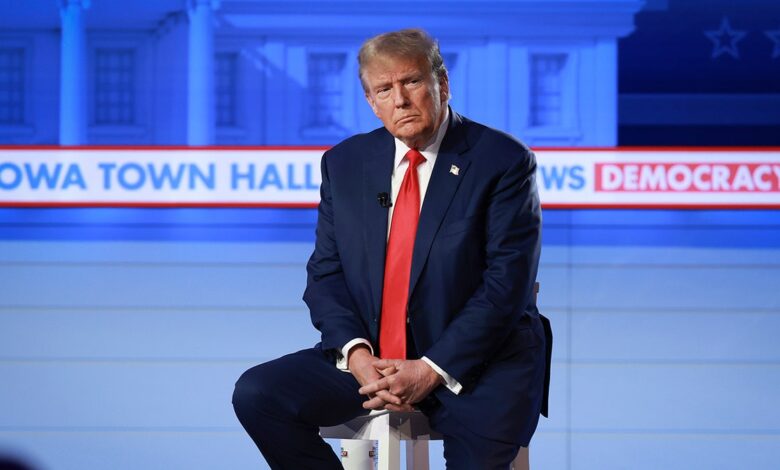 Trump with serious look on his face, sitting down on stool on stage, folded hands across lap, wearing a navy suit with a bright red tie