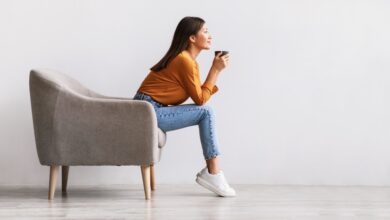 Young Asian woman sitting in armchair drinking hot coffee against a white wall