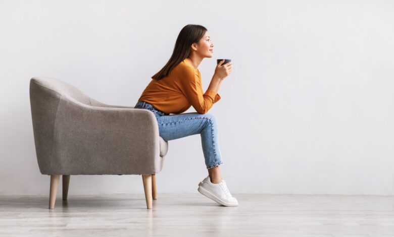 Young Asian woman sitting in armchair drinking hot coffee against a white wall