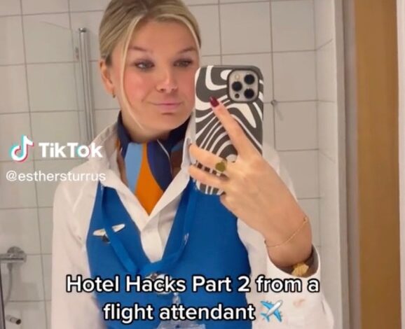 A flight attendant named Esther Sturrus taking a mirror selfie while demonstrating her hotel hack of rolling a water bottle under the bed for safety
