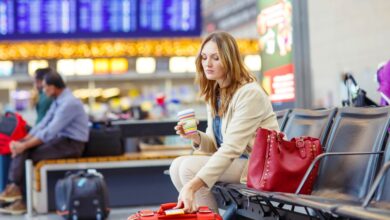 Woman sitting and drinking coffee at an international airport terminal with a red bag, looking upset due to a canceled flight.