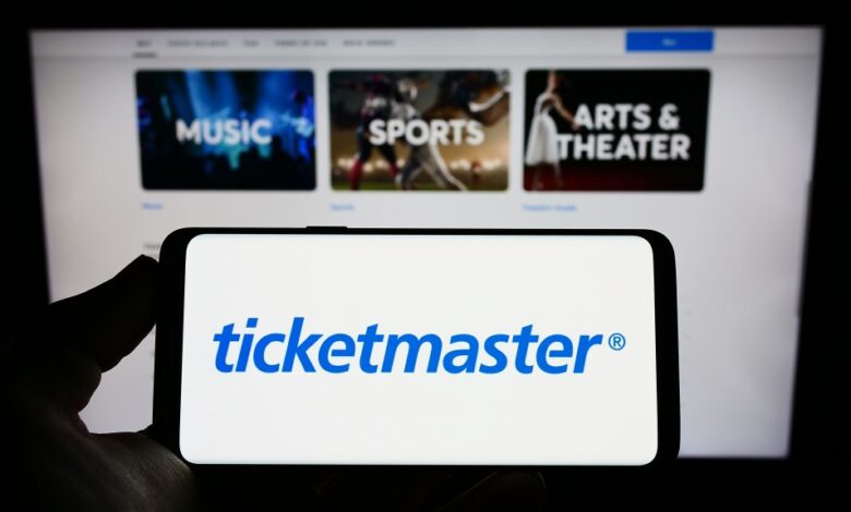 Person holding a cellphone displaying the logo of Ticketmaster Entertainment Inc, with a business webpage in the background.