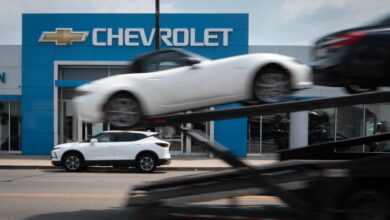 A car hauler passing a Chevrolet dealership in Chicago, representing the halted workflow due to a cyber attack on auto dealership software systems