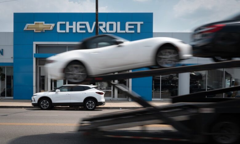 A car hauler passing a Chevrolet dealership in Chicago, representing the halted workflow due to a cyber attack on auto dealership software systems