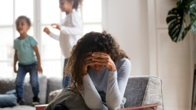 Black single frustrated woman hold her head with hands sitting on chair in living room, playful kids jumping on couch on a background. Tiredness, depression difficult to educate children alone concept