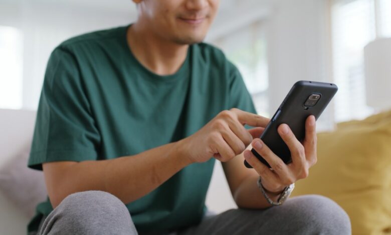 Adult Asian man sitting on sofa at home, smiling and swiping right on a dating app on his cell phone