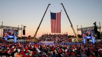 A crowd wearing red, white, and blue gathers at a Trump rally with two cranes holding up an American flag as the backdrop.