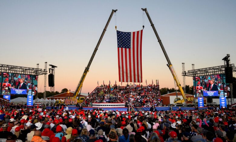 A crowd wearing red, white, and blue gathers at a Trump rally with two cranes holding up an American flag as the backdrop.