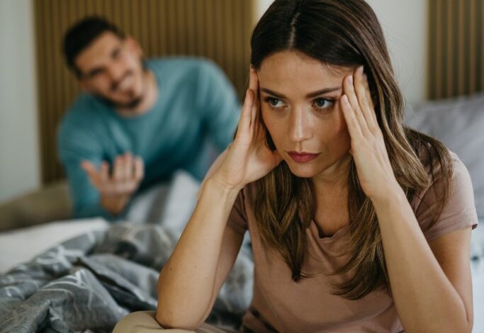 Young couple having marital problems in the bedroom, with upset woman on the bed and arguing husband in the background