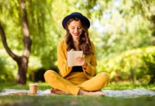 A cheerful, wavy-haired woman sitting in lotus position on a blanket outdoors, making notes