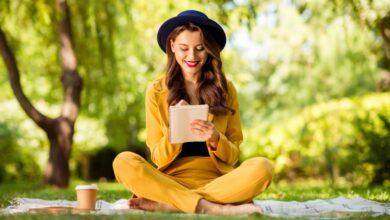 A cheerful, wavy-haired woman sitting in lotus position on a blanket outdoors, making notes