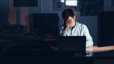 Young Asian businessman looking exhausted with his head in his hand while working alone at his desk in an office late at night