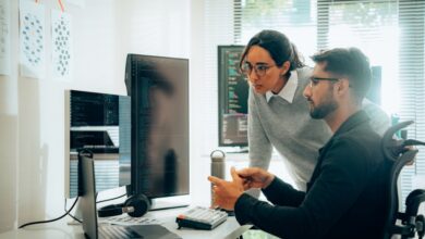 Two technology professionals work diligently in a bright office setting, one focusing on a laptop while the other engages with a large monitor, coding together.