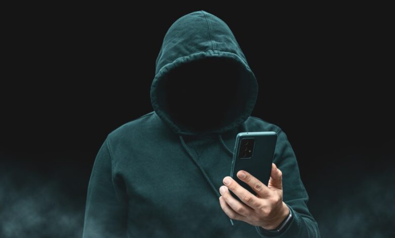 Silhouette of a faceless, hooded figure, presumably a hacker or cybercriminal, holding a smartphone