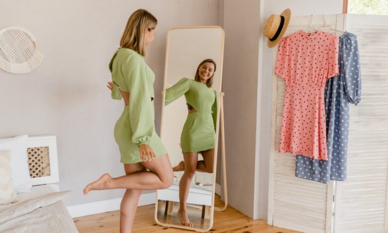 Young woman in a green dress trying on a stylish dress and looking in a mirror at home or a showroom