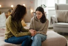 A mother and teenage daughter holding hands and talking on a bed.