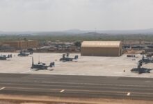 U.S. military aircraft, including MV-22 Ospreys, are parked on the tarmac at a forward operating base in Djibouti, with aircraft hangars and support structures visible under hazy skies near the Horn of Africa.
