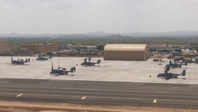U.S. military aircraft, including MV-22 Ospreys, are parked on the tarmac at a forward operating base in Djibouti, with aircraft hangars and support structures visible under hazy skies near the Horn of Africa.