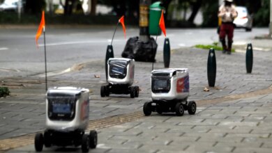 Three delivery robots with orange flags on a paved sidewalk next to a street.