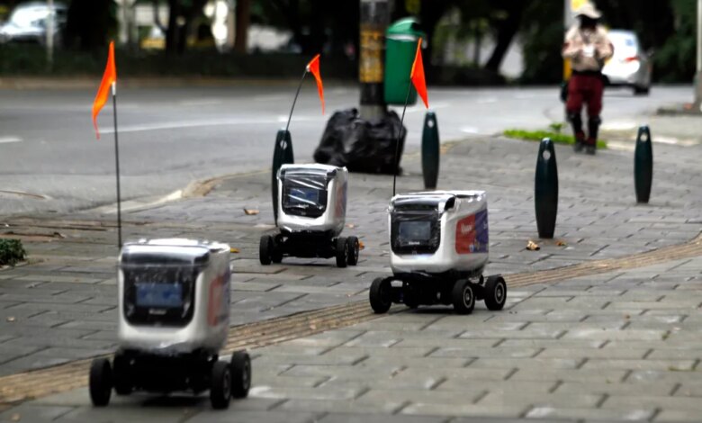 Three delivery robots with orange flags on a paved sidewalk next to a street.