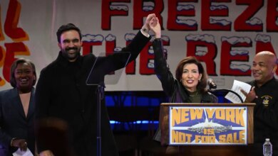 Zohran Mamdani and Kathy Hochul raise joined hands, with other politicians, in front of a "New York is Not For Sale" banner.