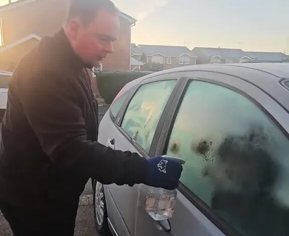 A man defrosts his car window with warm water in a spray bottle.