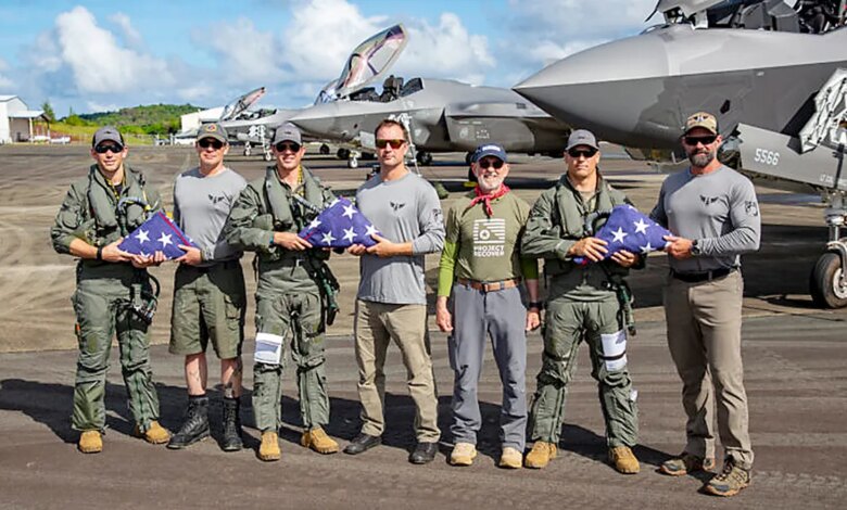 Men hold folded American flags on an airstrip beside military aircrafts.