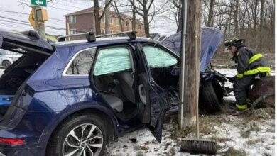 A blue SUV after a car crash, with air bags deployed and trunk open, and a firefighter inspecting the heavily damaged front of the car.