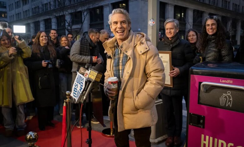 Andy Cohen smiles in a tan puffer coat holding a coffee cup, standing next to a hot pink "Landfill" trash can with "Hudson Square" written on its side.