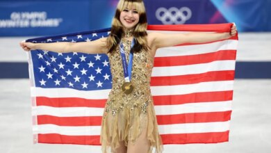Gold medalist Alysa Liu of Team United States, in a sparkling gold dress, holds an American flag behind her shoulders during the medal ceremony.