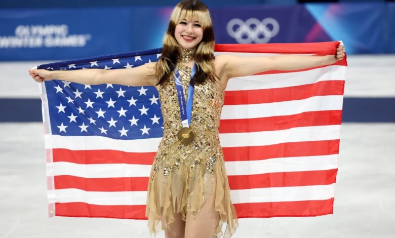 Gold medalist Alysa Liu of Team United States, in a sparkling gold dress, holds an American flag behind her shoulders during the medal ceremony.