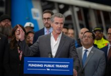 California Governor Gavin Newsom speaking at a podium with “Funding Public Transit” written on it, with other people in the background.