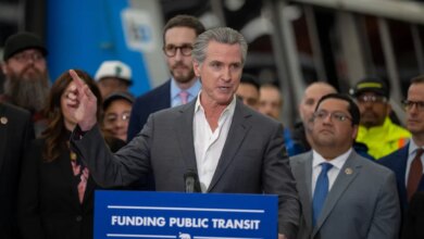 California Governor Gavin Newsom speaking at a podium with “Funding Public Transit” written on it, with other people in the background.