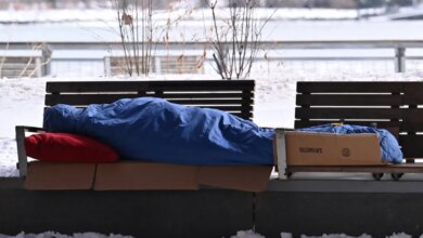 A homeless person sleeping on a park bench covered in a blue tarp with a red pillow, surrounded by snow.
