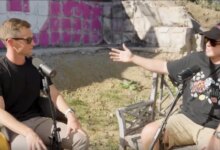 Two men in sunglasses having a discussion with microphones outdoors.