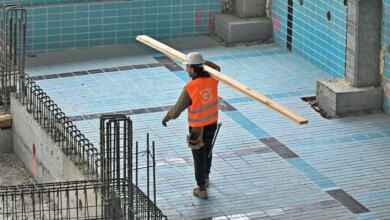 A construction worker in a hard hat and safety vest carries a wooden plank across a partially tiled swimming pool undergoing renovation.