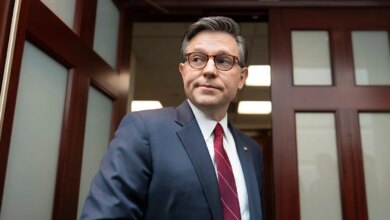 Speaker Mike Johnson standing still and looking toward reporters in a hallway at the Capitol.