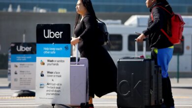 Two women with luggage wait for an Uber at Los Angeles International Airport, next to a sign with COVID-19 safety guidelines.