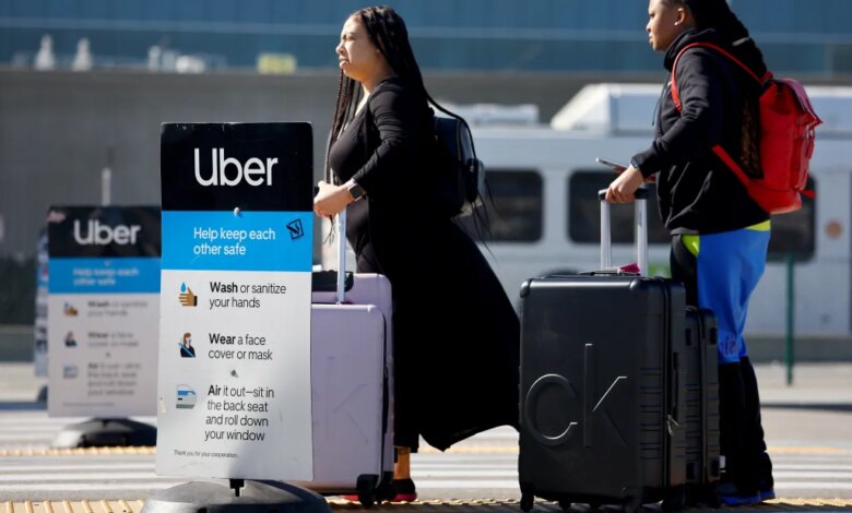 Two women with luggage wait for an Uber at Los Angeles International Airport, next to a sign with COVID-19 safety guidelines.