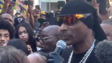 Snoop Dogg wearing a black du-rag and sunglasses, speaking into a microphone.