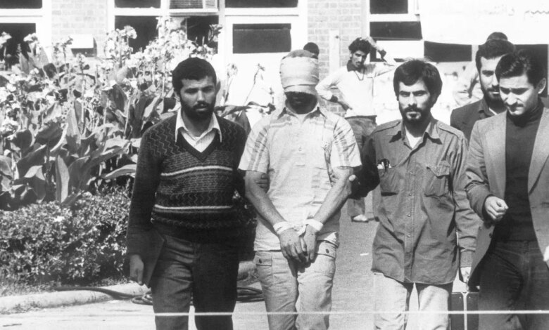 A blindfolded and bound hostage is led by Iranian students in front of a building.