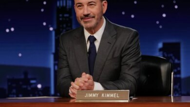 TV host Jimmy Kimmel at his desk in a dark suit, tie, and white shirt, smiling slightly.