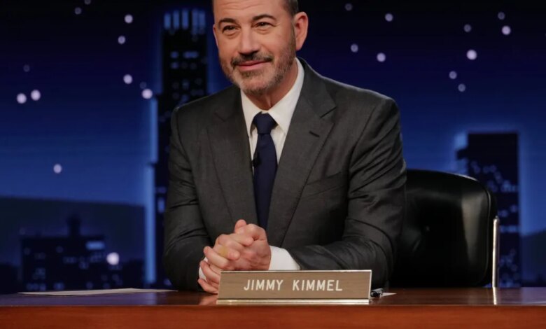 TV host Jimmy Kimmel at his desk in a dark suit, tie, and white shirt, smiling slightly.
