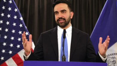 Mayor Zohran Mamdani speaking at a press conference with American and New York flags in the background.