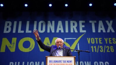 Senator Bernie Sanders speaks at a podium in front of a blue banner that reads "Billionaire Tax Now!" and "Vote Yes 11/3/26".