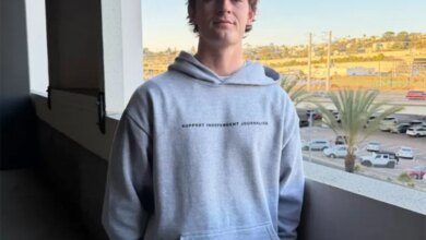 A young man in a grey hoodie with "SUPPORT INDEPENDENT JOURNALISM" printed on it stands on a balcony overlooking a city.