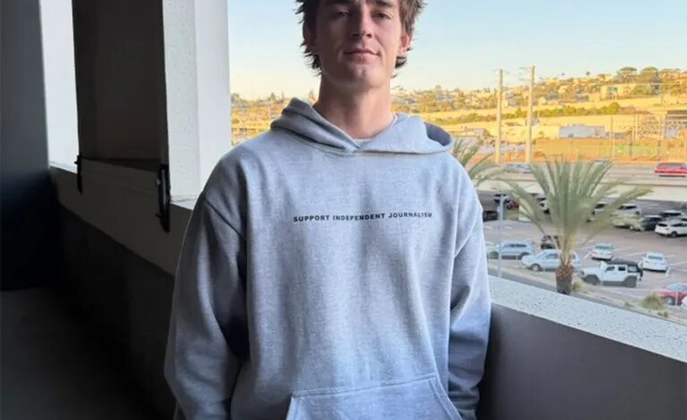A young man in a grey hoodie with "SUPPORT INDEPENDENT JOURNALISM" printed on it stands on a balcony overlooking a city.