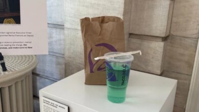 A Taco Bell Baja Blast drink with a straw, a brown Taco Bell paper bag, and a plaque about "Delivering Relief for Workers and Small Businesses" on a white pedestal in what appears to be a museum exhibit.